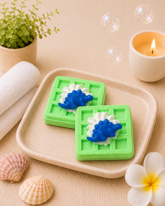 Two green waffle-shaped soap bars with blue and white decorations on a wooden tray, surrounded by bubbles, shells, and a candle.