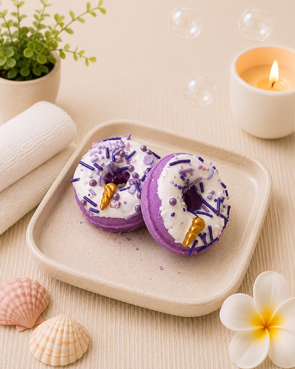 Two purple bath bombs with gold accents on a beige tray, surrounded by a candle, plant, and seashells.