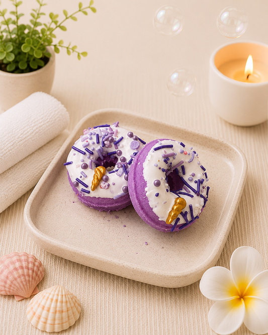 Two purple bath bombs with gold accents on a beige tray, surrounded by a candle, plant, and seashells.