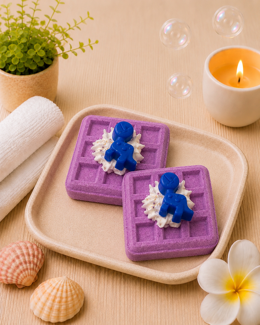 Two purple waffle-shaped soap bars with blue figures on a beige tray, surrounded by bubbles, a candle, and decorative items.