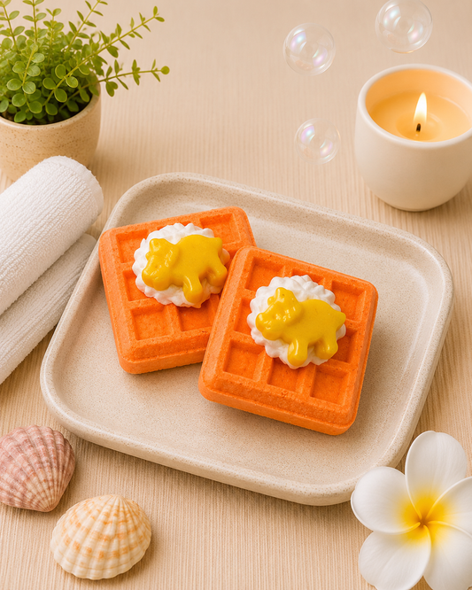 Two orange waffle-shaped soap bars with yellow centers on a beige plate, surrounded by bubbles, a candle, shells, and a plant.