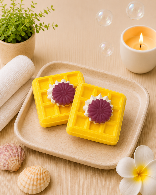 Two yellow soap bars with purple flowers on a beige plate, surrounded by bubbles and a candle.